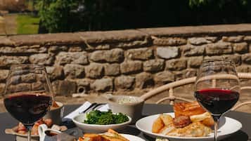 Una mesa servida con platos de comida, dos copas de vino tinto y una silla de mimbre en un jardín.
