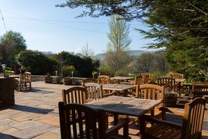 Un patio al aire libre con mesas y sillas de madera, suelo de piedra y plantas en macetas.