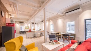 A spacious living area with a yellow armchair, red sofa, and a coffee table with a floral arrangement. There is a kitchen area in the background with a bar and stools. - Nornyaowarat Hostel (Bangkok)