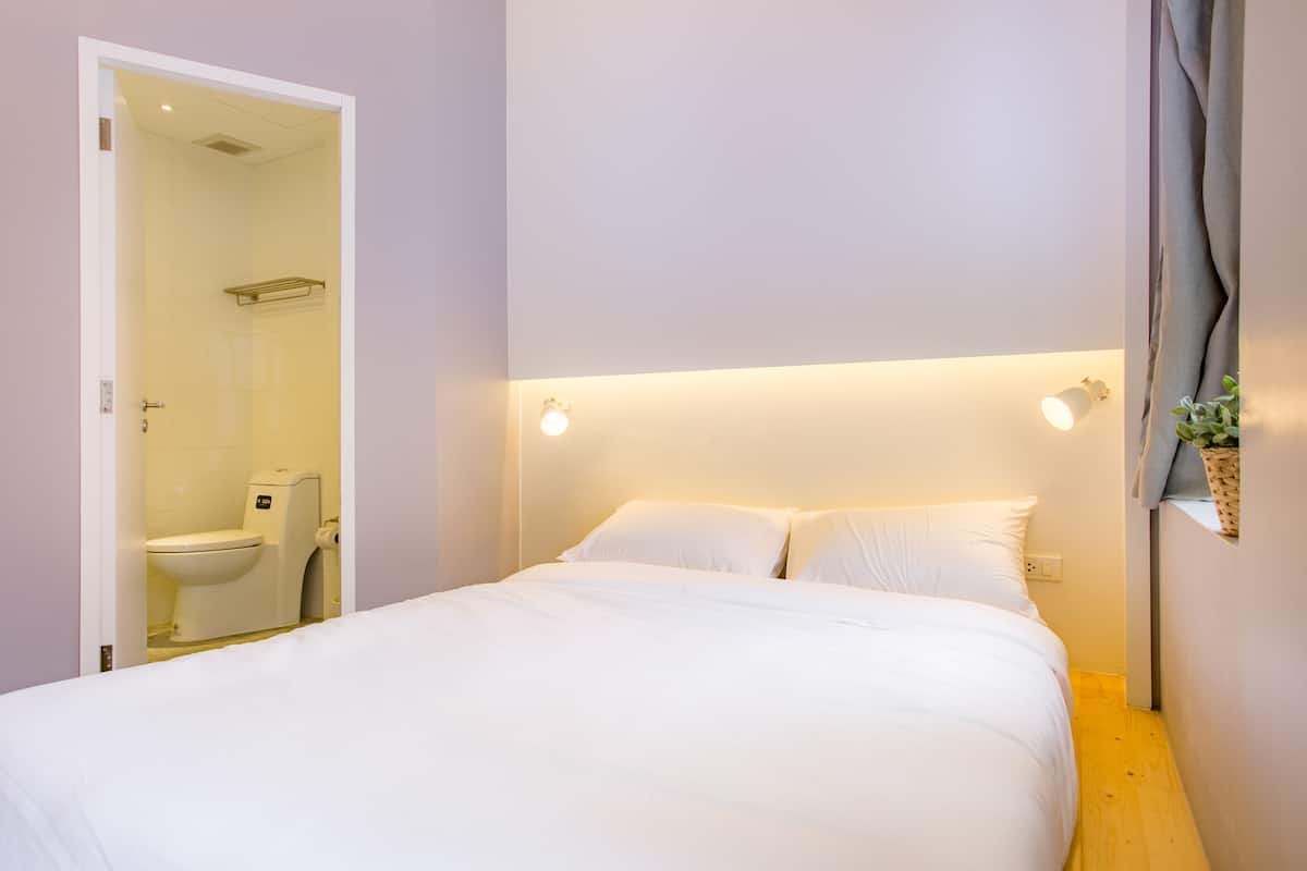 A hotel room with a bed, a bedside table, a nightstand, a wall-mounted light, and a window with curtains.
