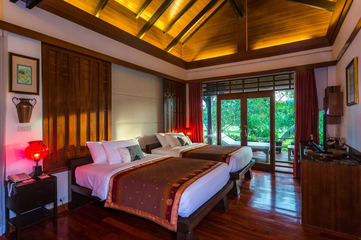 Une chambre d’hôtel avec deux lits, un plancher en bois, un plafond avec des poutres apparentes, une table de chevet avec une lampe et une fenêtre donnant sur un paysage verdoyant.