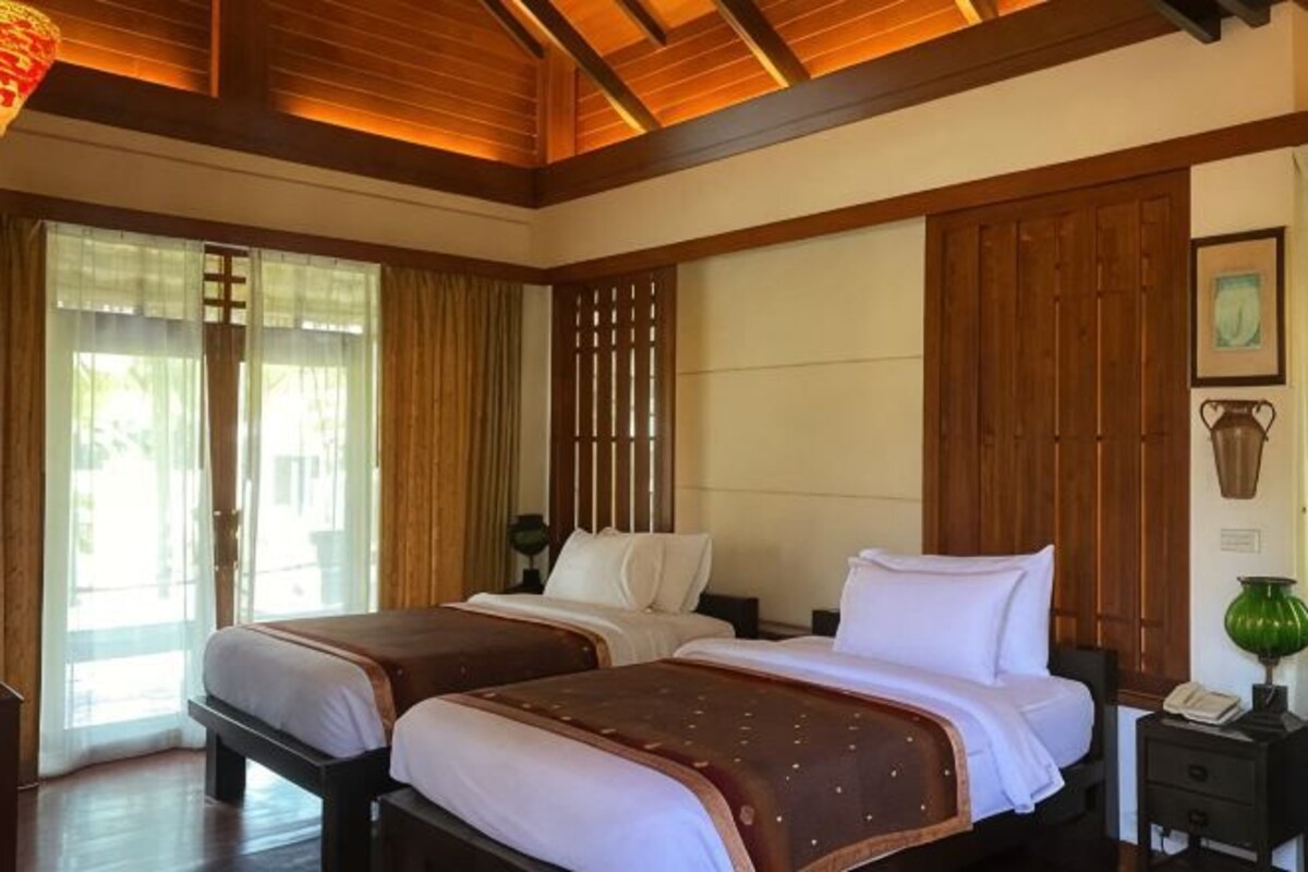 Een hotelkamer met twee bedden, een houten plafond, een groot raam met gordijnen en een groene vaas op een nachtkastje.