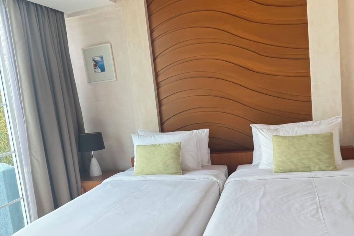 Un lit double avec du linge de lit blanc et deux oreillers, une tête de lit en bois, une table de chevet avec une lampe et une fenêtre avec des rideaux.