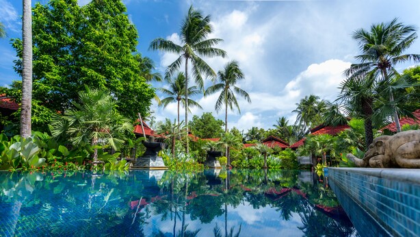 A clear pool surrounded by lush greenery and palm trees, with a stone sculpture on the right.