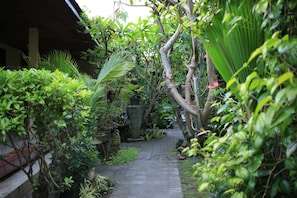 A narrow stone pathway flanked by lush greenery and potted plants, leading to a covered area with a wooden structure. - Simon Homestay (Denpasar)