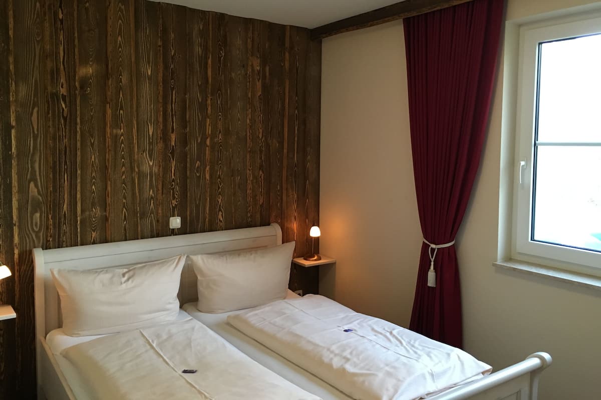 En dobbeltseng med hvide sengetøj, en træpanelfront, et natbord med lampe, og et vindue med røde gardiner.