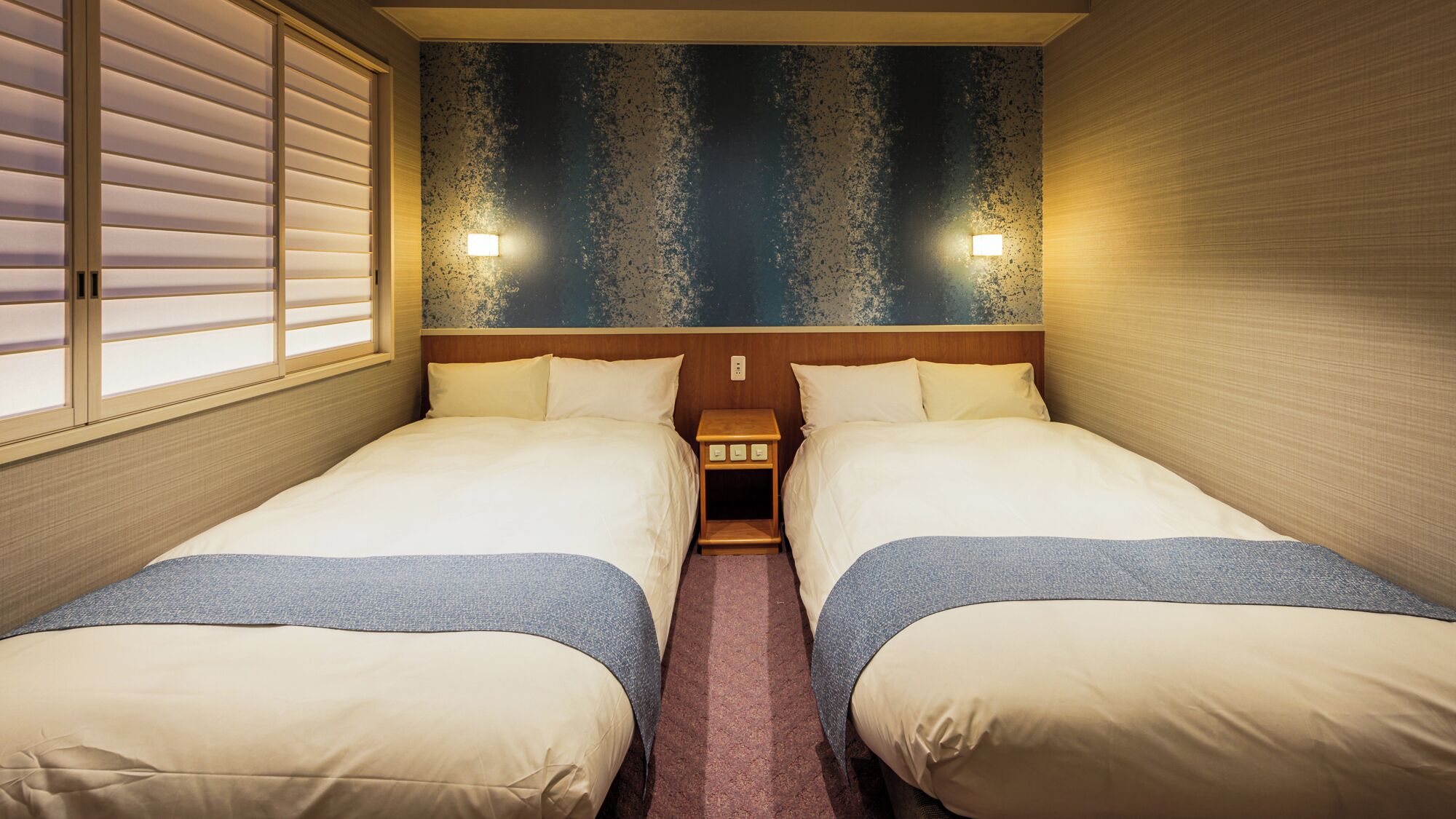 A double room with two beds, a small bedside table with a lamp, and a textured wall.