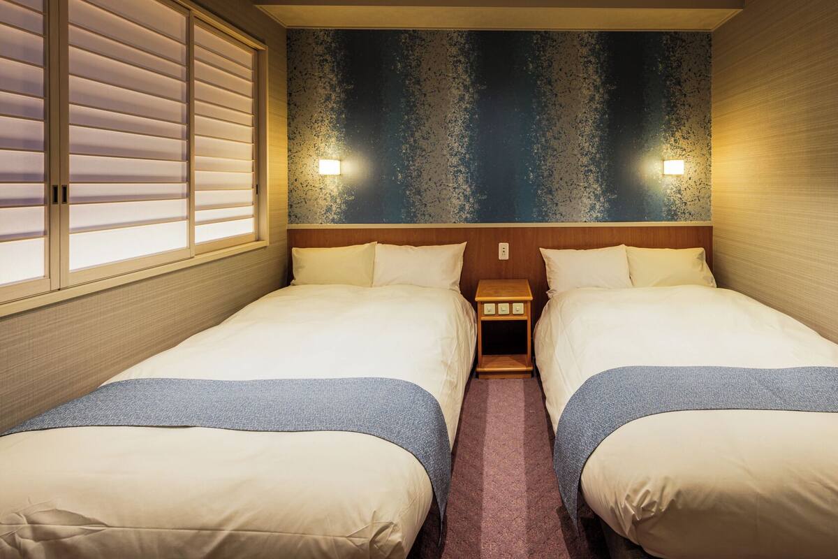 A double room with two beds, a small bedside table with a lamp, and a textured wall.