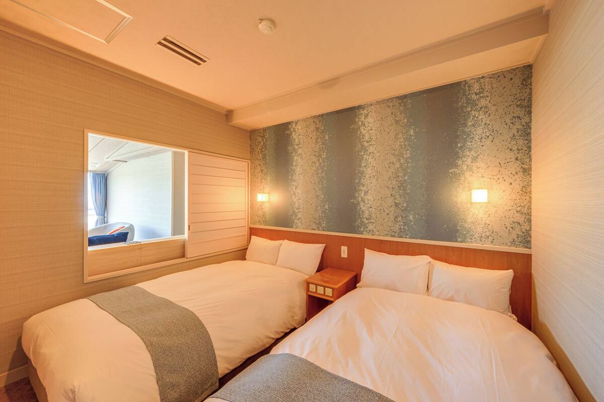A hotel room with two beds, a large window, a textured wall, and a nightstand.
