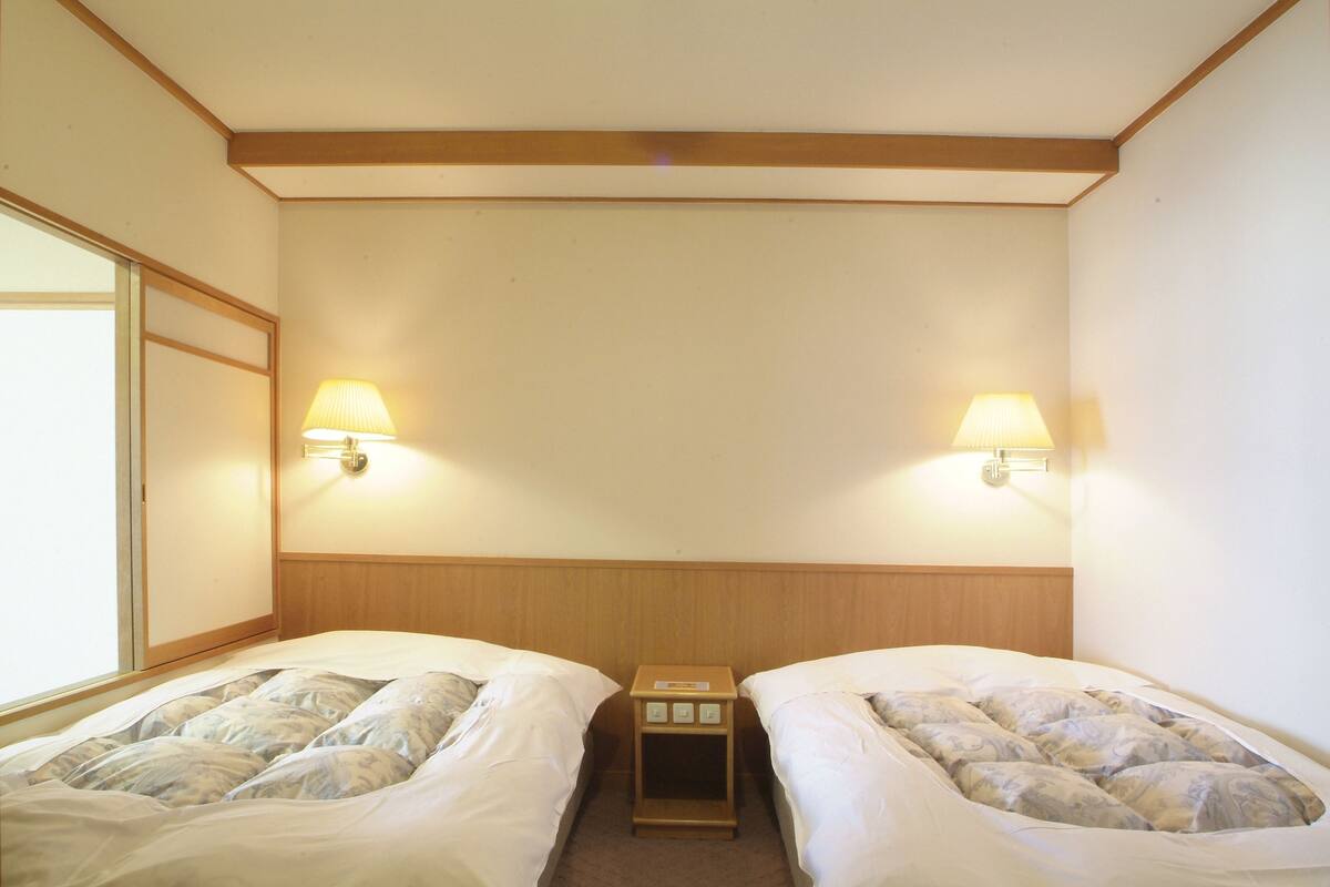 A hotel room with two single beds, each with a pillow and a blanket. There are two wall-mounted lamps on either side of the bed.