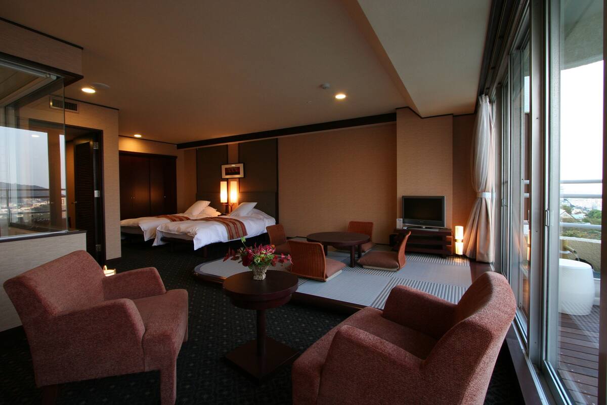 A hotel room with a bed, two armchairs, a small table with a vase, a TV, and a large window offering a view.