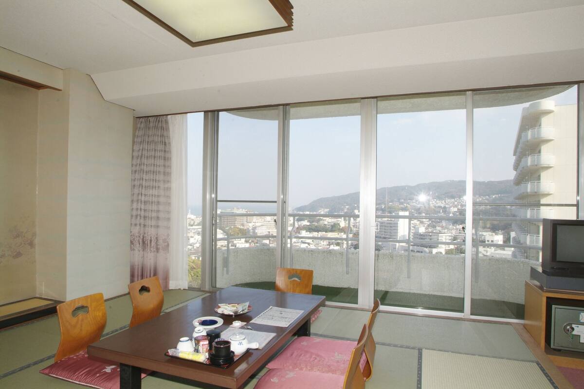 A room with a dining table, chairs, a TV, and a large window with a city view.