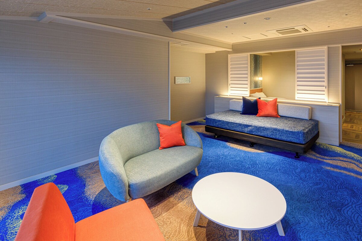 A hotel room with a sofa, a small table, a bed with pillows, and a mirror.
