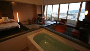 A hotel room with a bed, a sitting area with a table and chairs, a large window with a city view, and a bathtub. - Hotel Juraku (Ito)