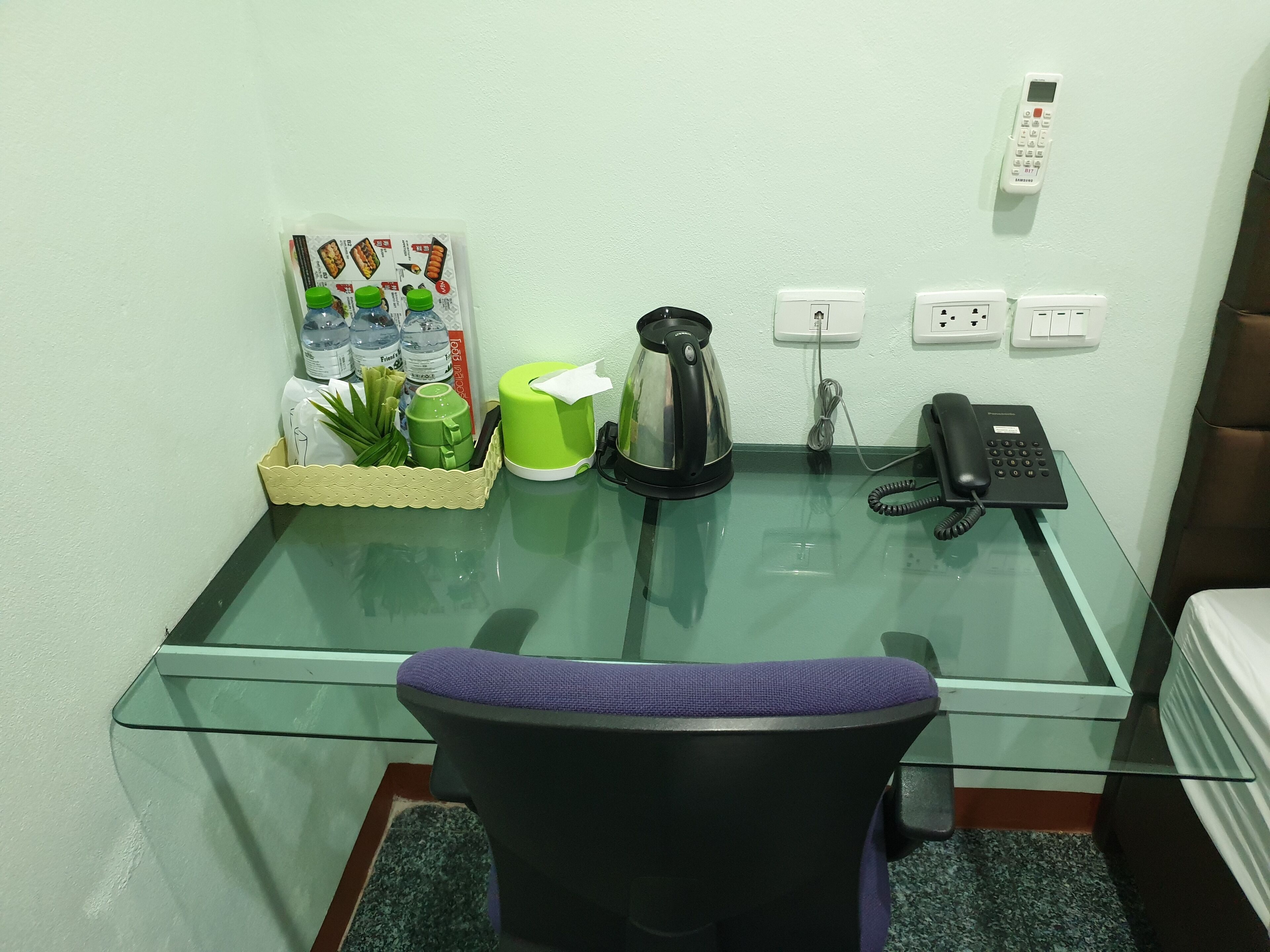Un bureau à plateau en verre, une chaise de bureau noire, une cafetière verte et blanche, un téléphone noir et un téléphone Samsung fixé au mur.