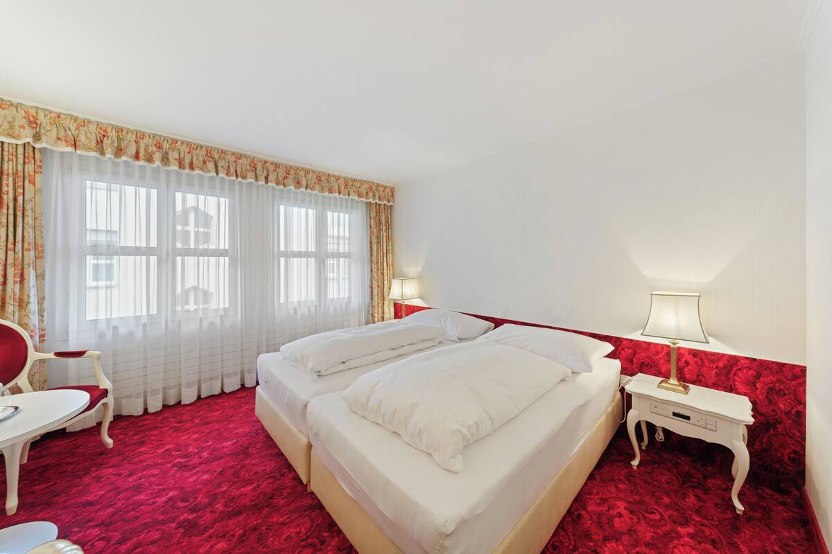 Quarto de hotel com duas camas, um tapete vermelho, uma cadeira branca, uma mesinha e uma mesa de cabeceira com abajur.