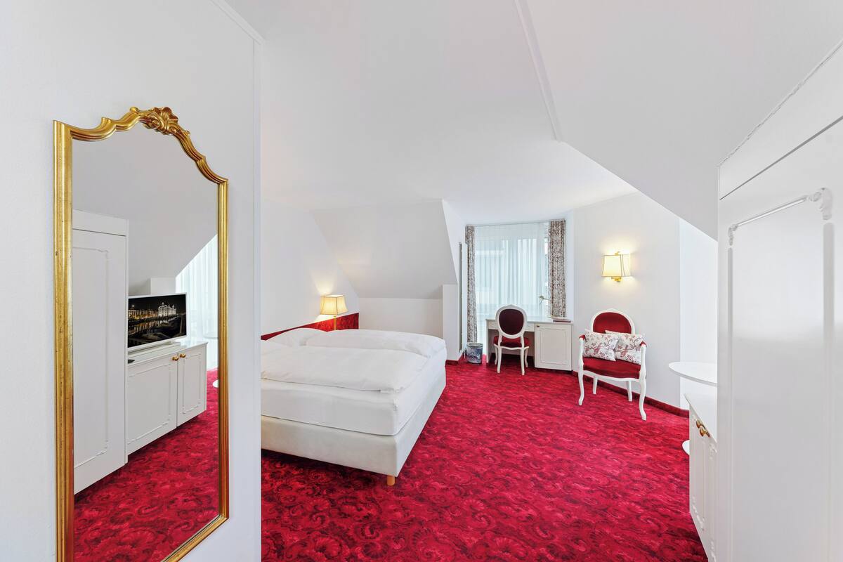 Quarto de hotel com carpete vermelho, mobiliário branco, um espelho grande, uma cama, uma escrivaninha e uma cadeira.