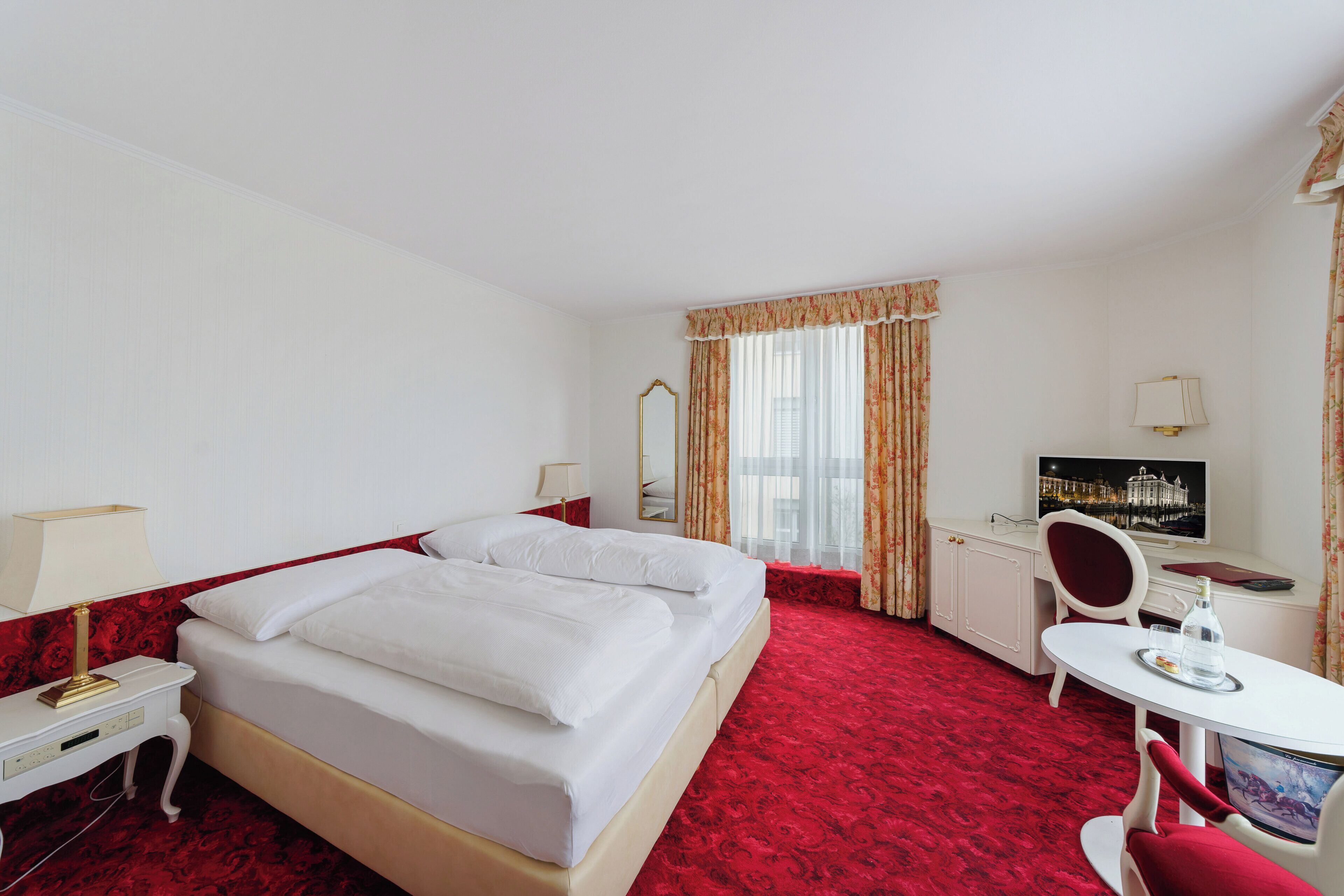 A hotel room with a bed, desk, chair, TV, and a red carpet.