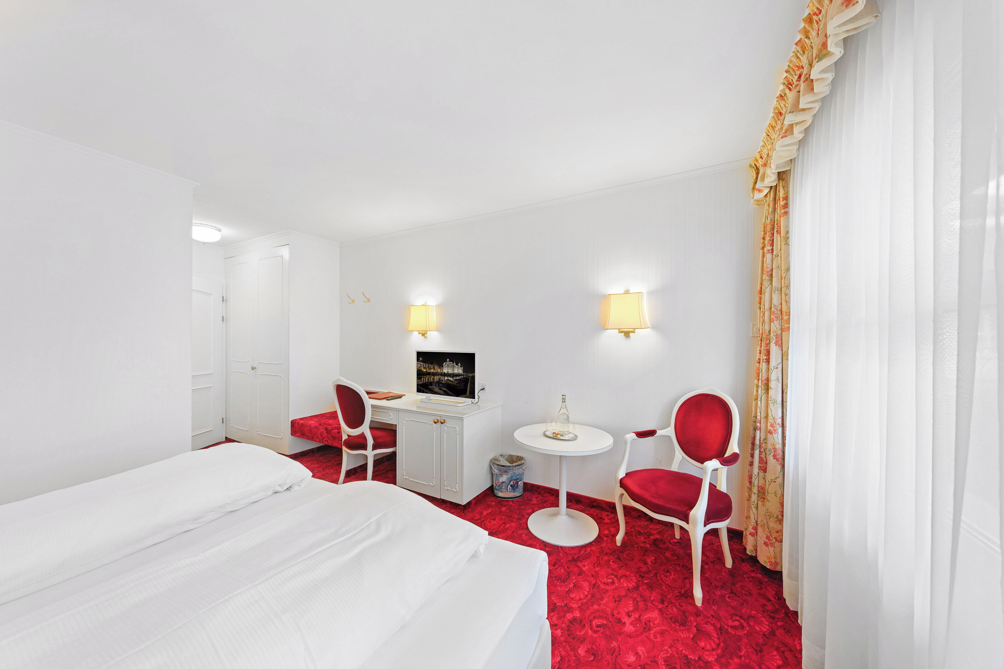 Habitación de hotel con cama, escritorio con computadora, dos sillas, una mesita y alfombra roja.