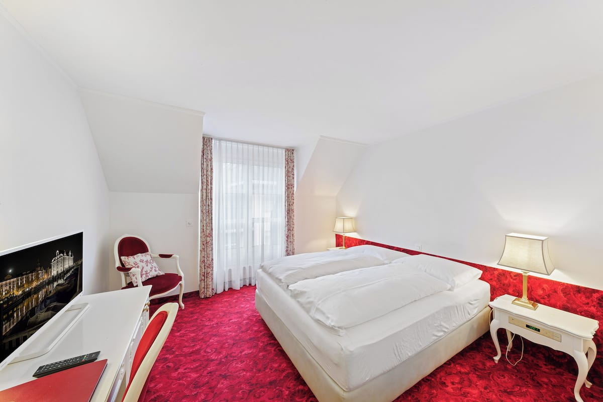 Quarto de hotel com cama, escrivaninha, cadeira, televisão e janela com cortinas.