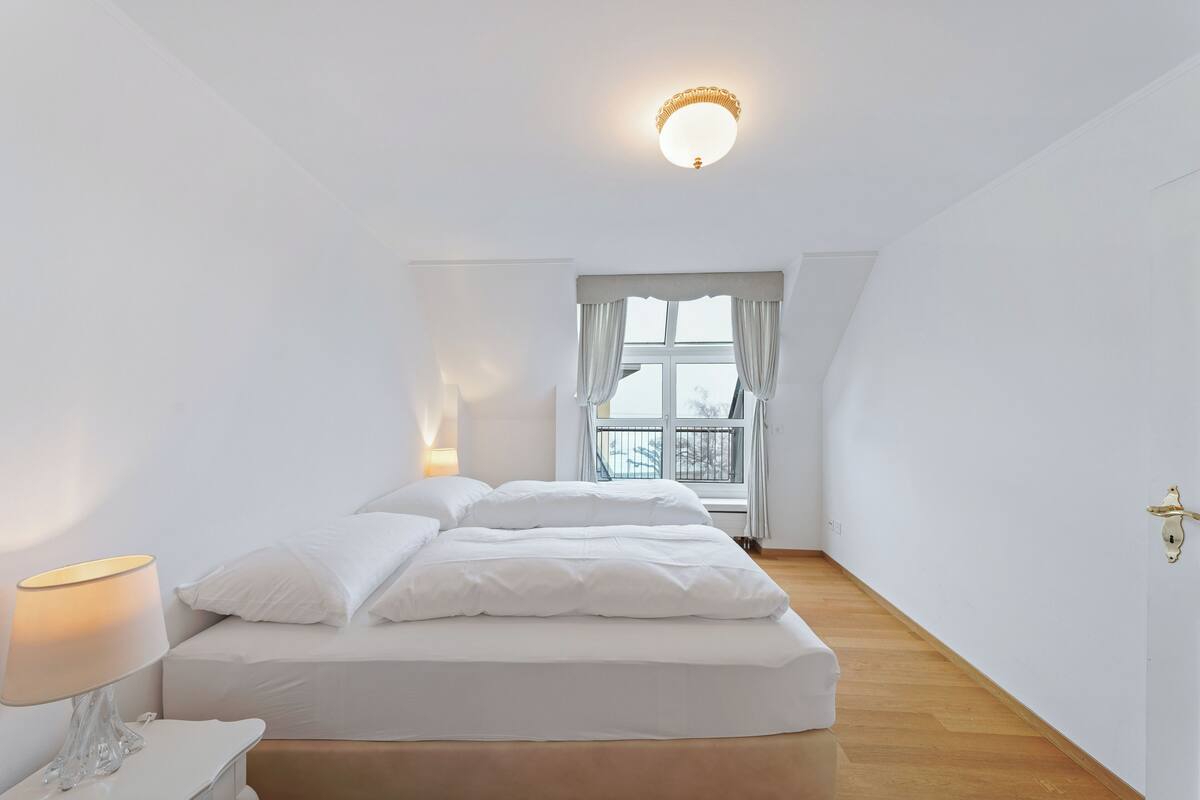 Quarto com cama, mesa de cabeceira, luminária, janela com cortinas e luminária de teto.