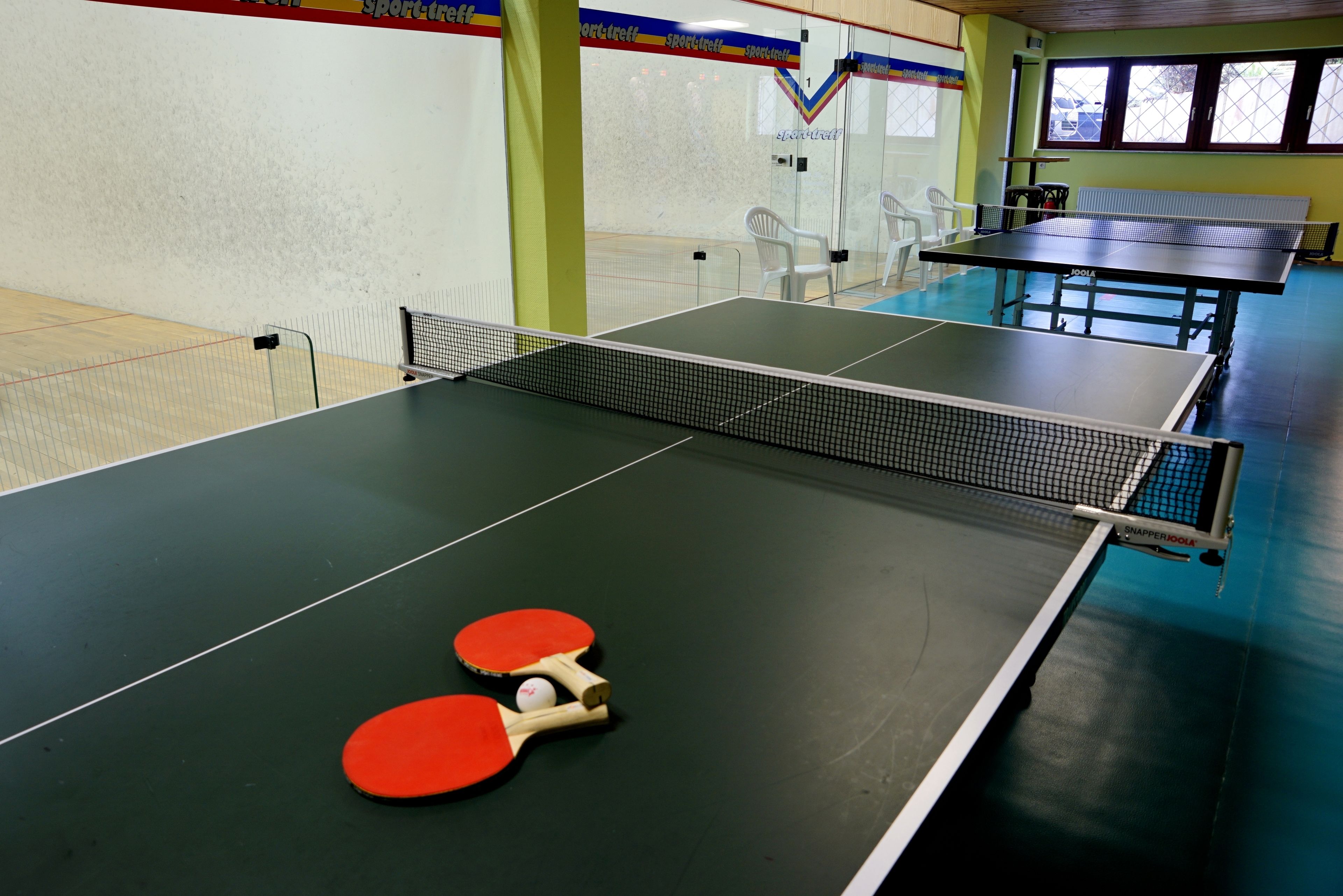 Una mesa de tenis de mesa con dos paletas rojas y una pelota, rodeada de sillas blancas y una pared verde con un cartel de temática deportiva.