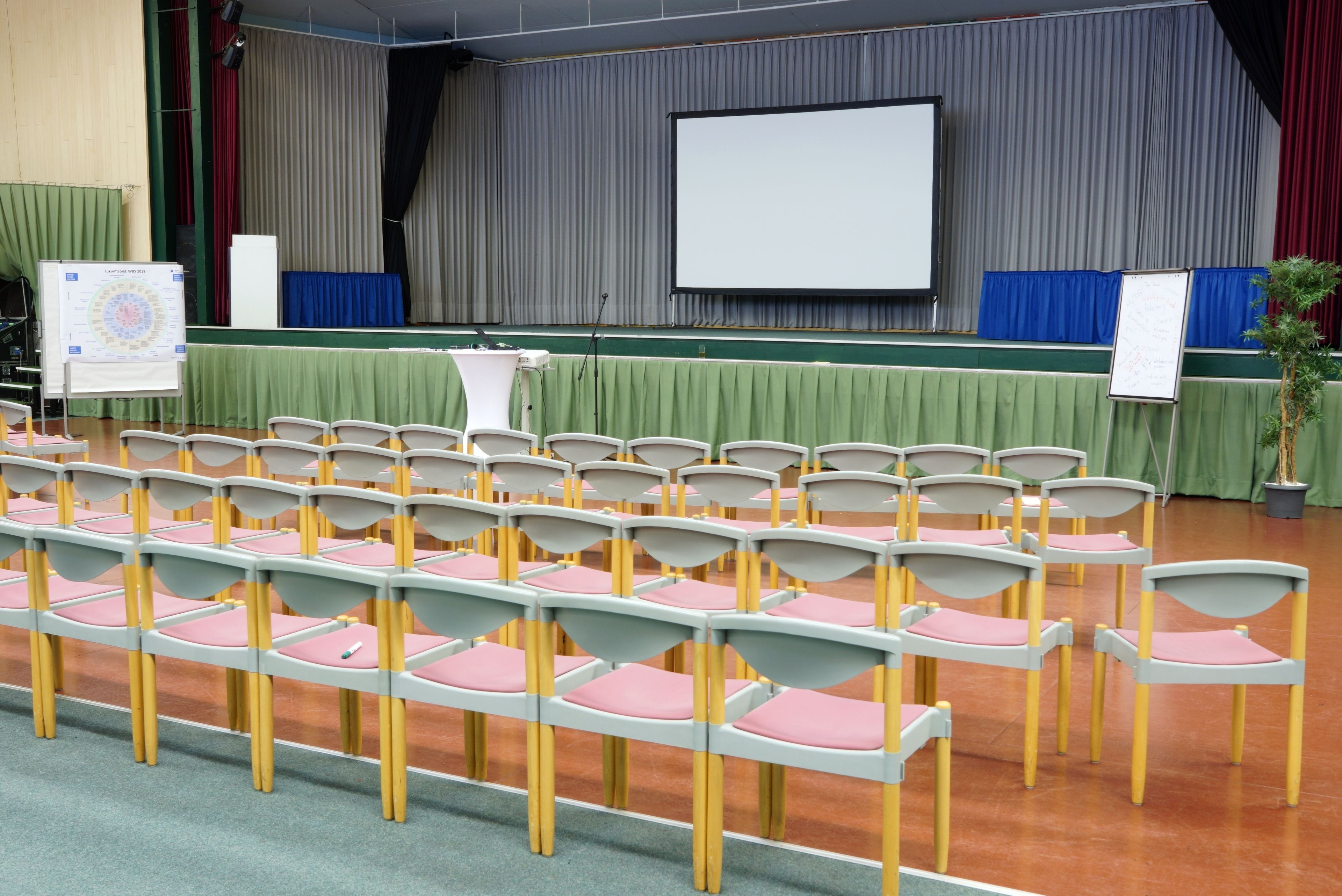 Un salón de conferencias con filas de sillas, un escenario con una pantalla y dos pizarras blancas.