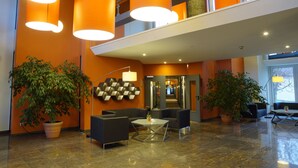 A modern lobby with orange walls, large hanging lights, potted plants, and contemporary furniture. - Trans World Hotel Auefeld (Hann. Muenden)