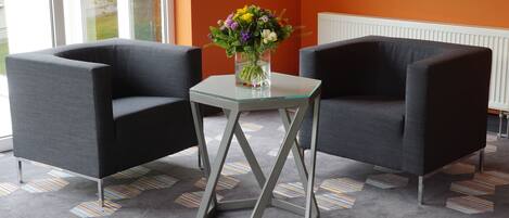 A room with two grey armchairs, a small glass table with a vase of flowers, and a patterned carpet.