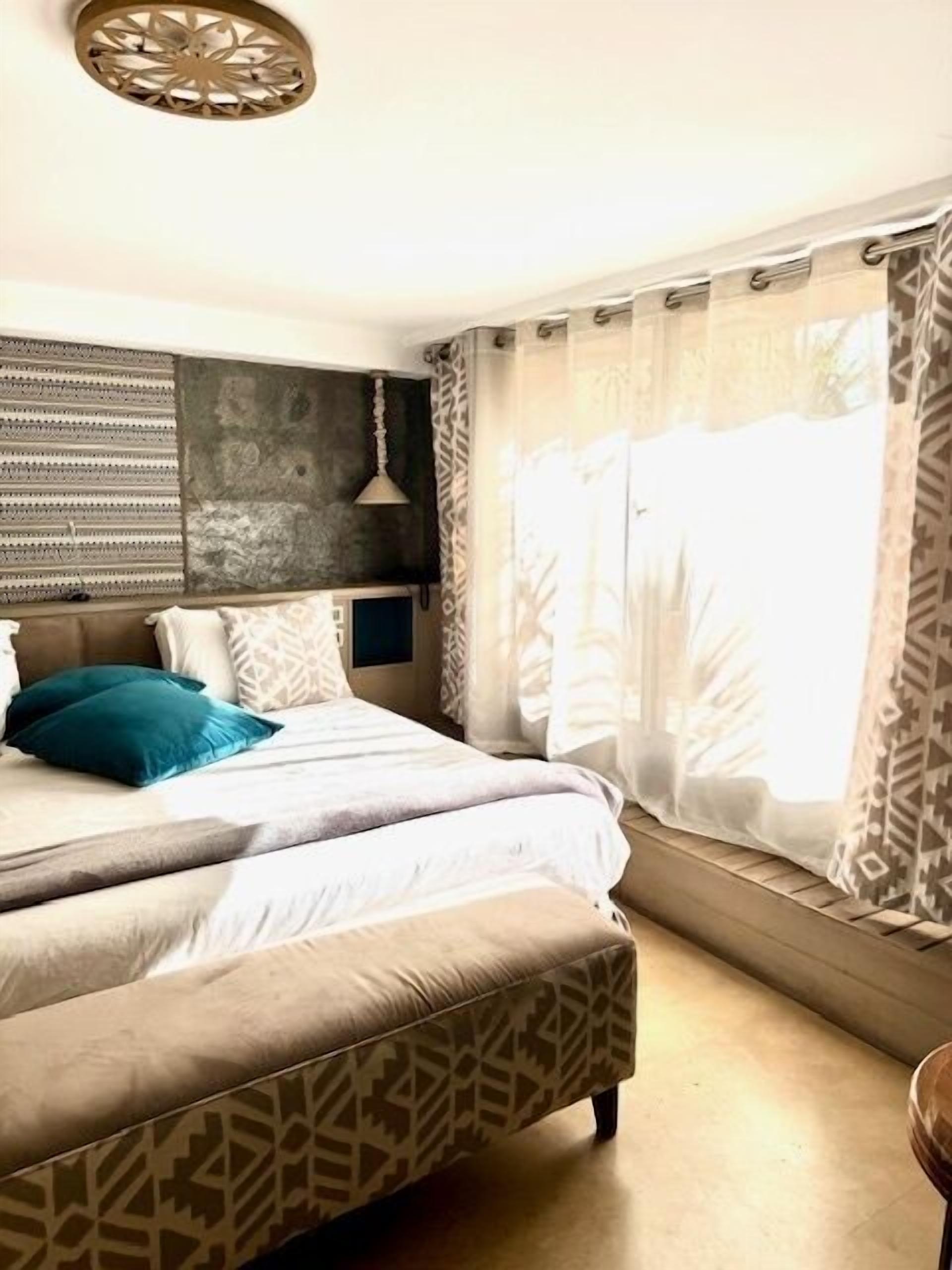 A bedroom with a large bed, a patterned ottoman, a hanging light fixture, and a window with sheer curtains.