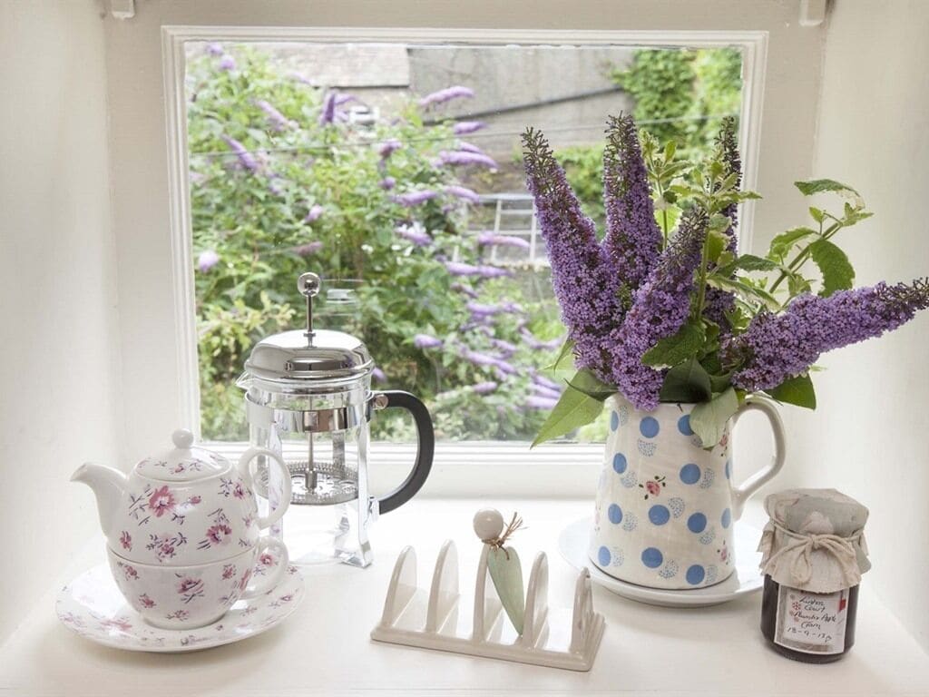 Blick aus dem Fenster auf einen weißen Teekessel, eine transparente Kaffeekanne, eine tischdurchmusterte Tasse und ein kleines Glas auf einem Fensterbrett.