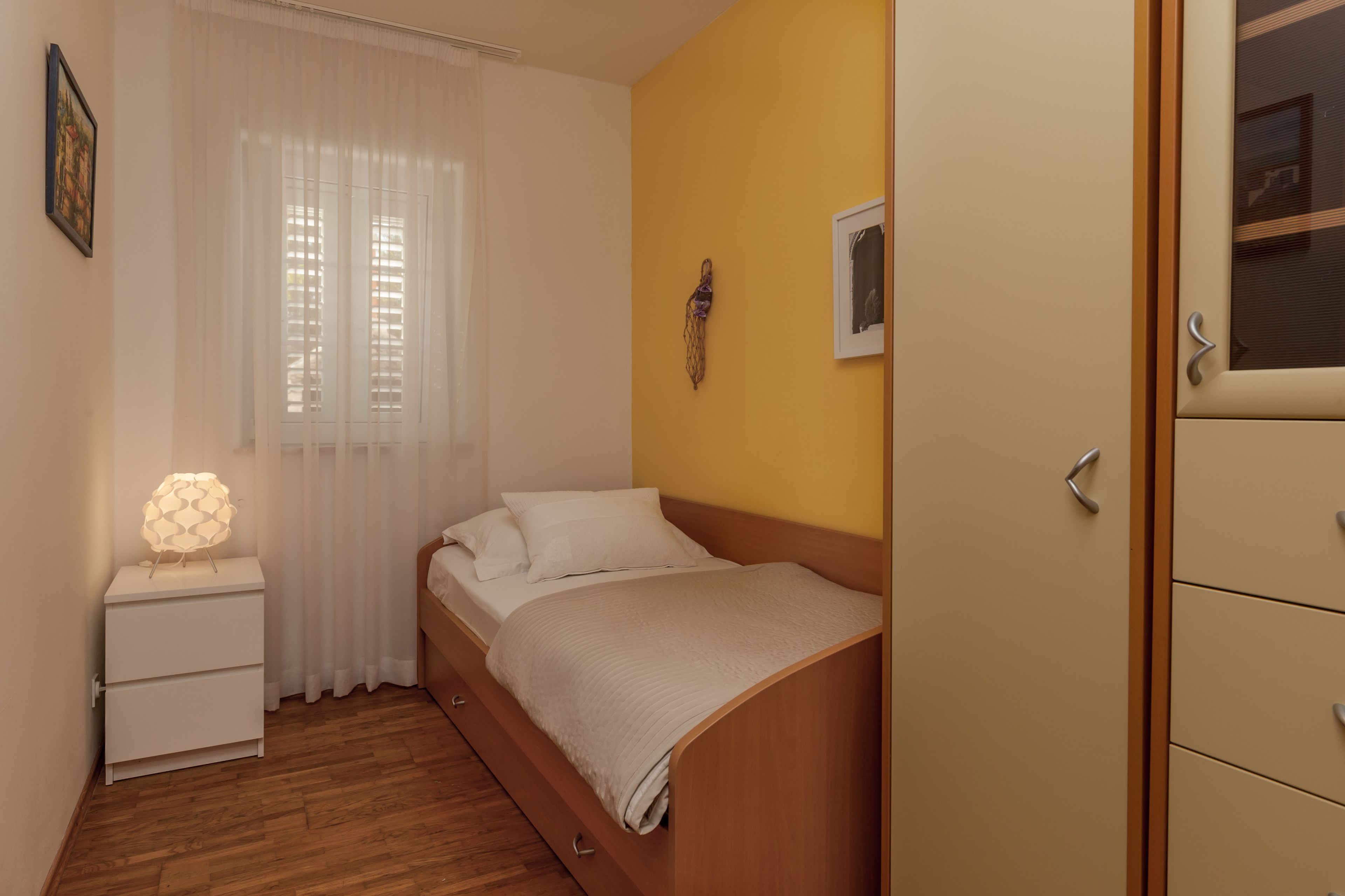 Un dormitorio con una cama de madera, una mesita de noche con lámpara, una ventana con cortinas translúcidas y una pared amarilla.