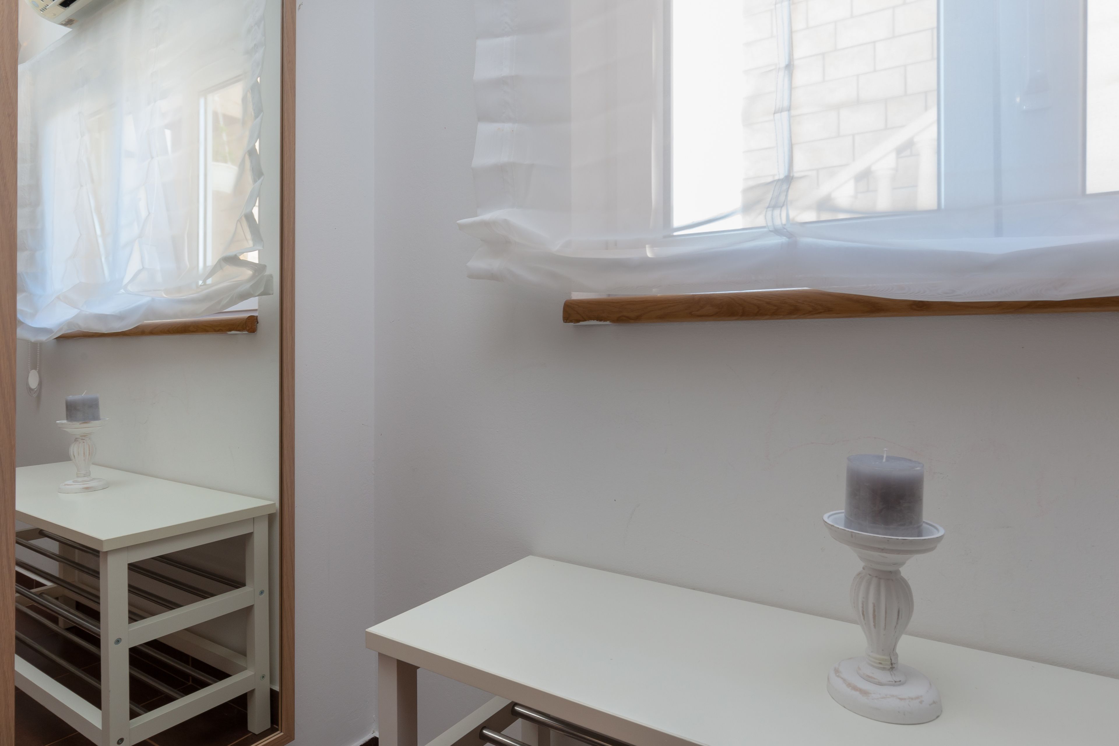 Una habitación con una mesa blanca, una vela sobre un atril, una ventana con cortinas traslúcidas y una pared de ladrillo afuera.