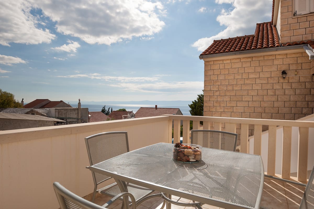 A balcony with a table and chairs, a view of the sea, and a brick building.