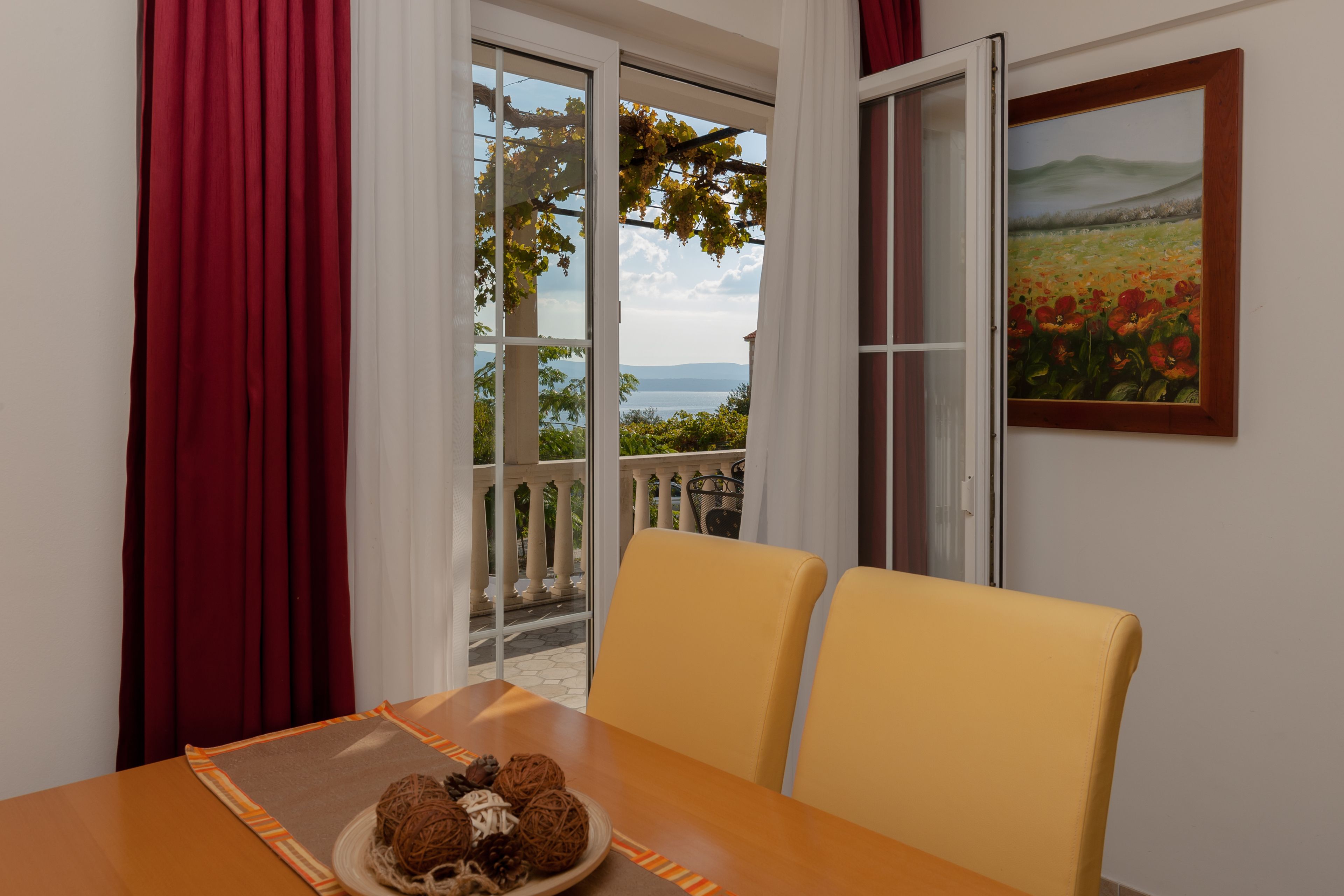 A dining area with a table set for two, a painting on the wall, and a view of greenery and a body of water through an open door.