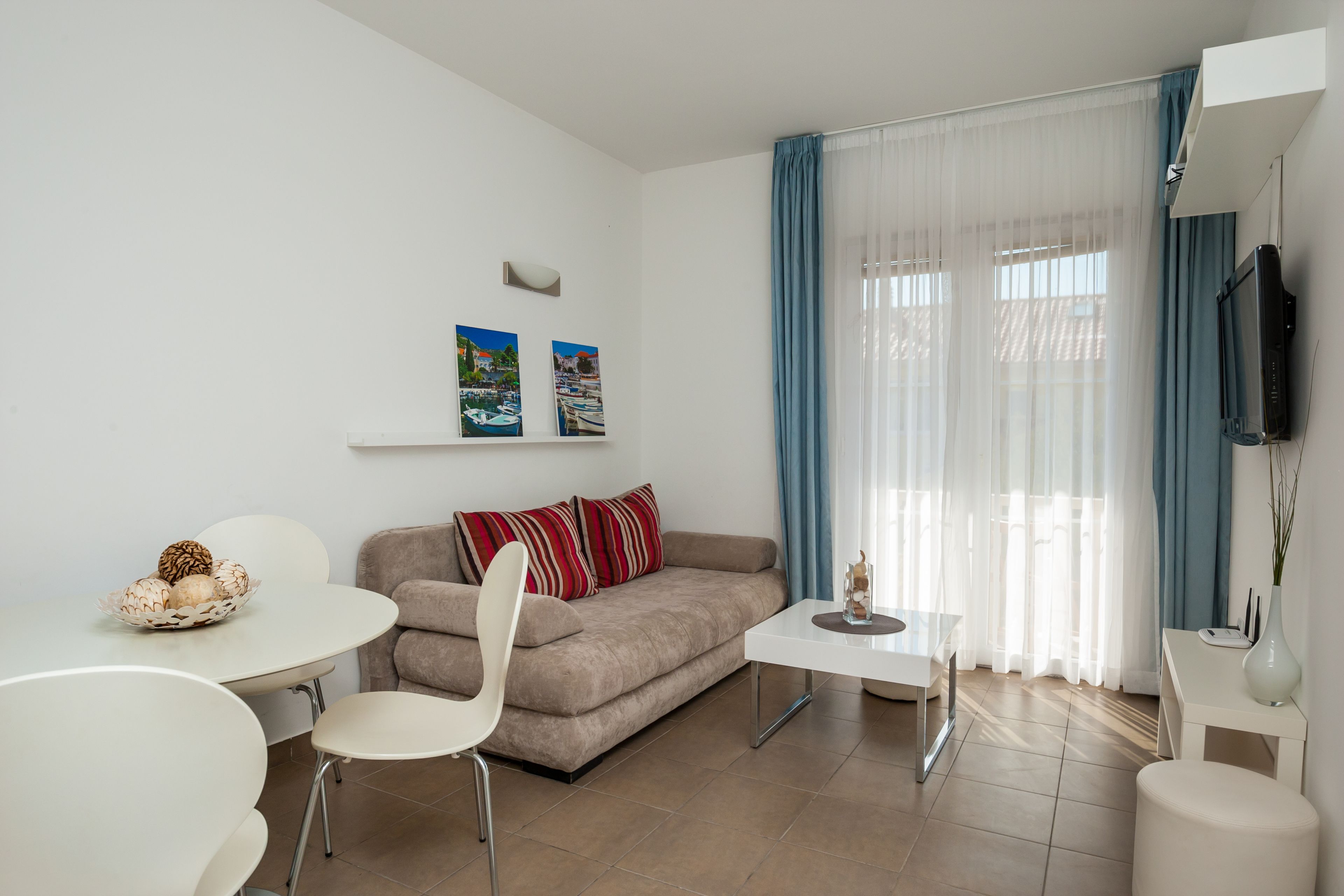 Una sala de estar moderna con un sofá color beige, sillas blancas, una pequeña mesa de comedor y un televisor montado en la pared.