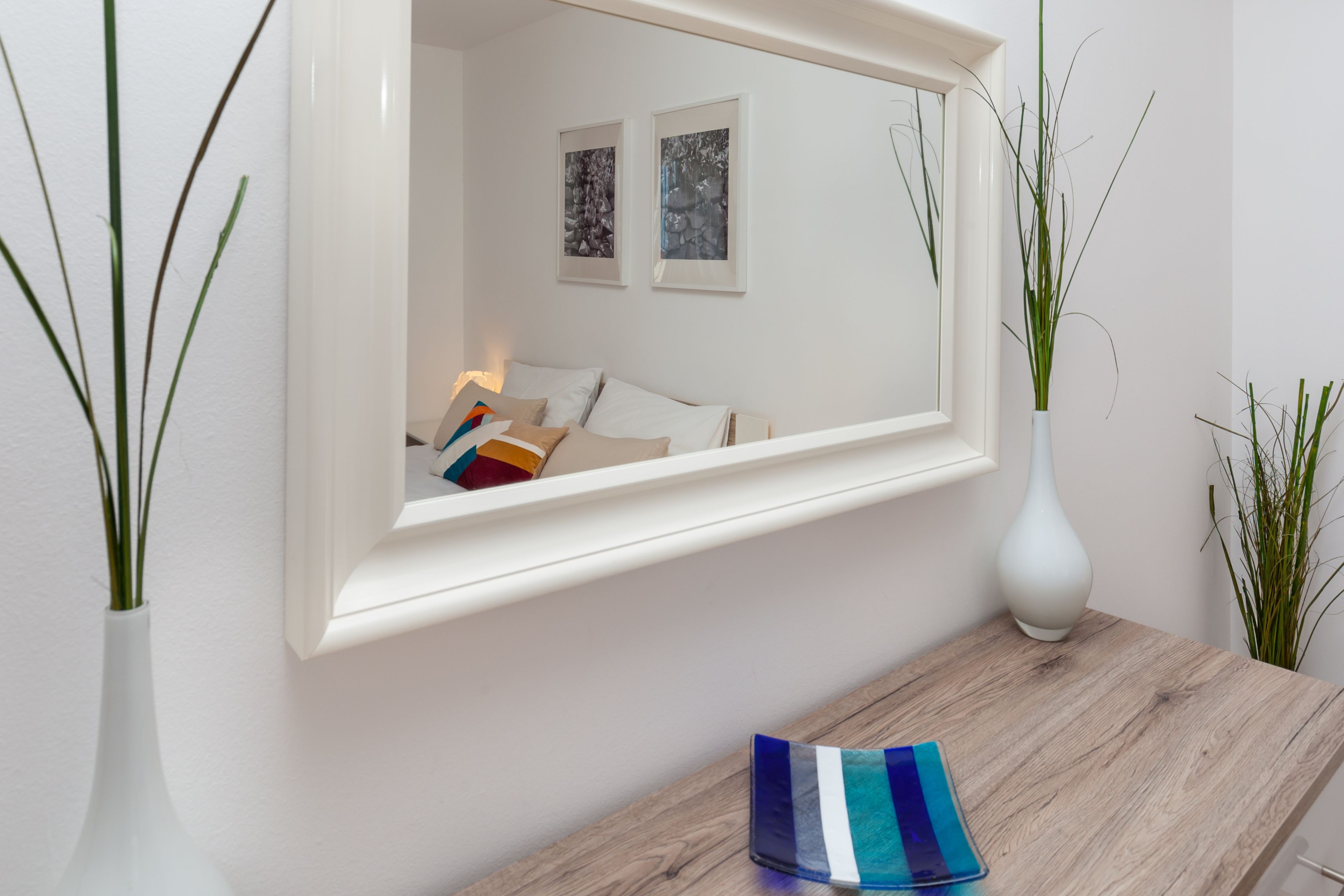 Una habitación con un espejo grande que refleja una cama, una mesa de madera y dos jarrones con plantas.