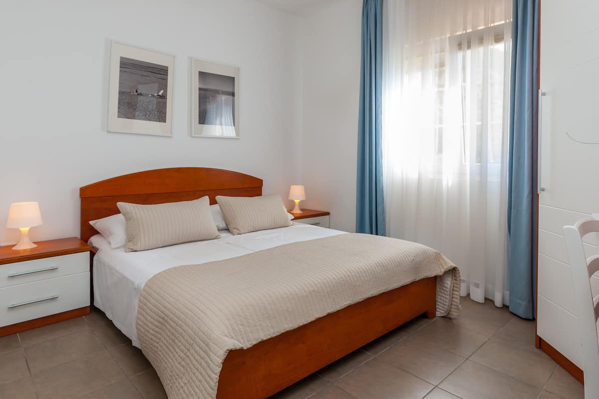 A bedroom with a wooden bed, white bedding, two bedside tables with lamps, and a window with curtains.