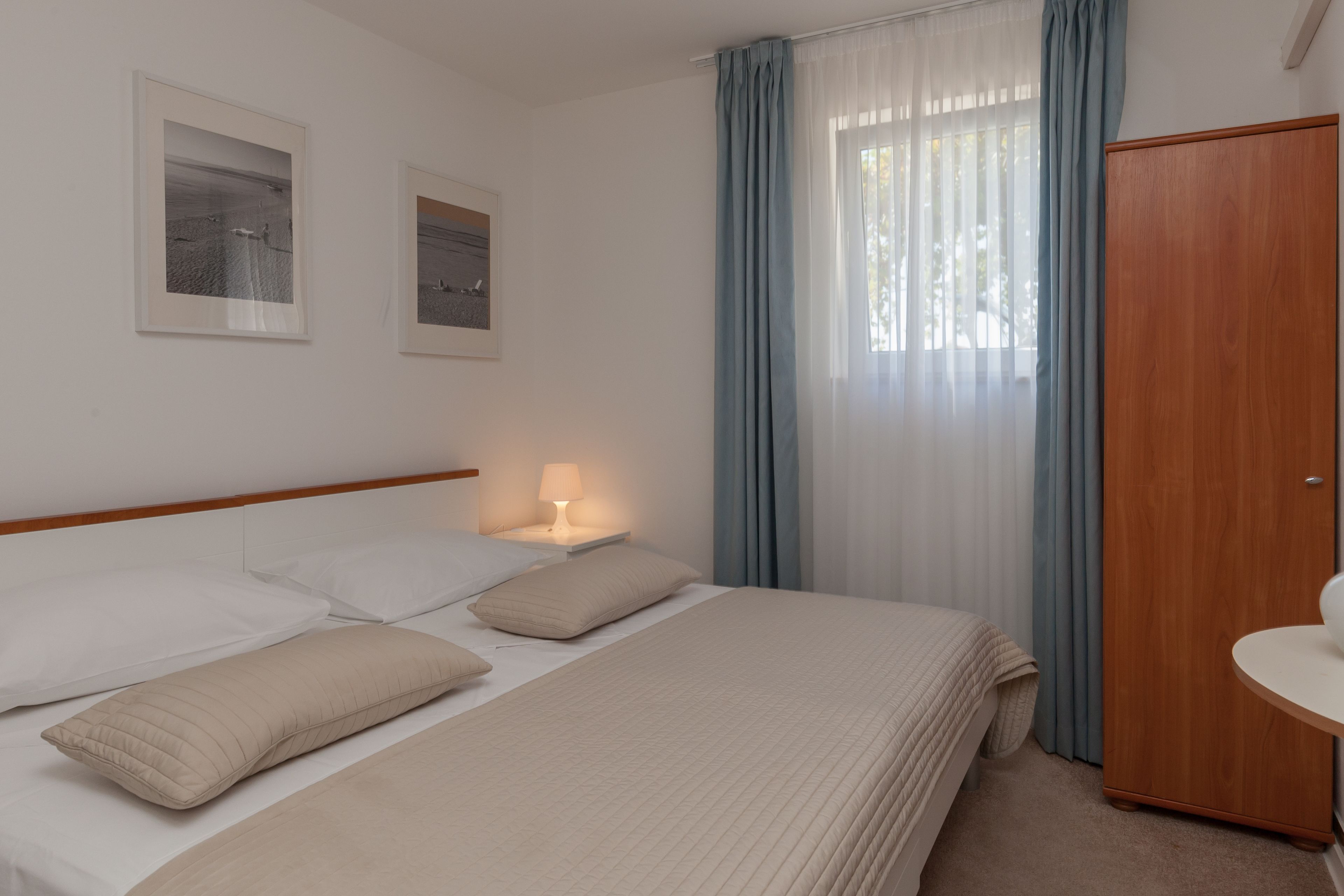 Cama bem arrumada com lençóis brancos e almofadas bege, um abajur, um guarda-roupa de madeira e duas fotos emolduradas na parede.