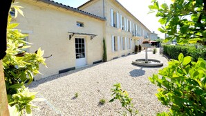 Une cour avec un chemin de gravier, une fontaine et un bâtiment doté d’un toit en tuiles et de fenêtres à cadre blanc.