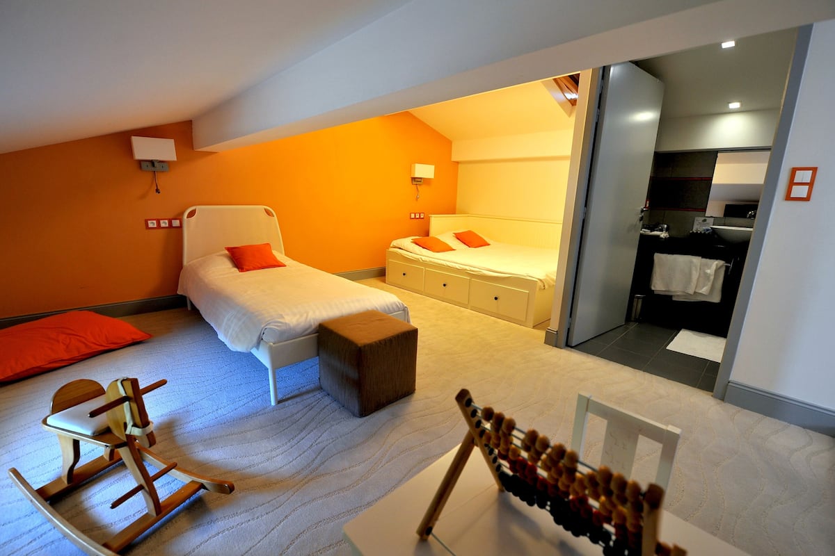 Une chambre d’hôtel avec deux lits, un fauteuil à bascule en bois, un petit tabouret et une plante suspendue.
