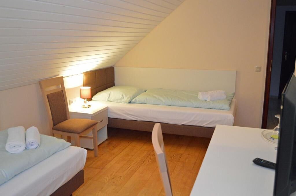 A small, cozy room with a sloped ceiling, a single bed, a bedside table with a lamp, a chair, and a TV.