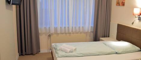 Habitación de hotel con cama, televisión montada en la pared, ventana con cortinas y lámpara de noche.
