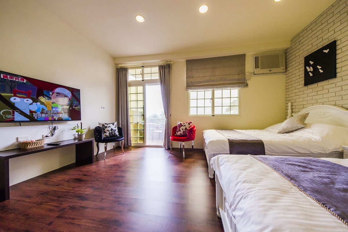Quarto de hotel com duas camas, uma televisão na parede esquerda, uma cadeira vermelha e uma janela com cortinas.