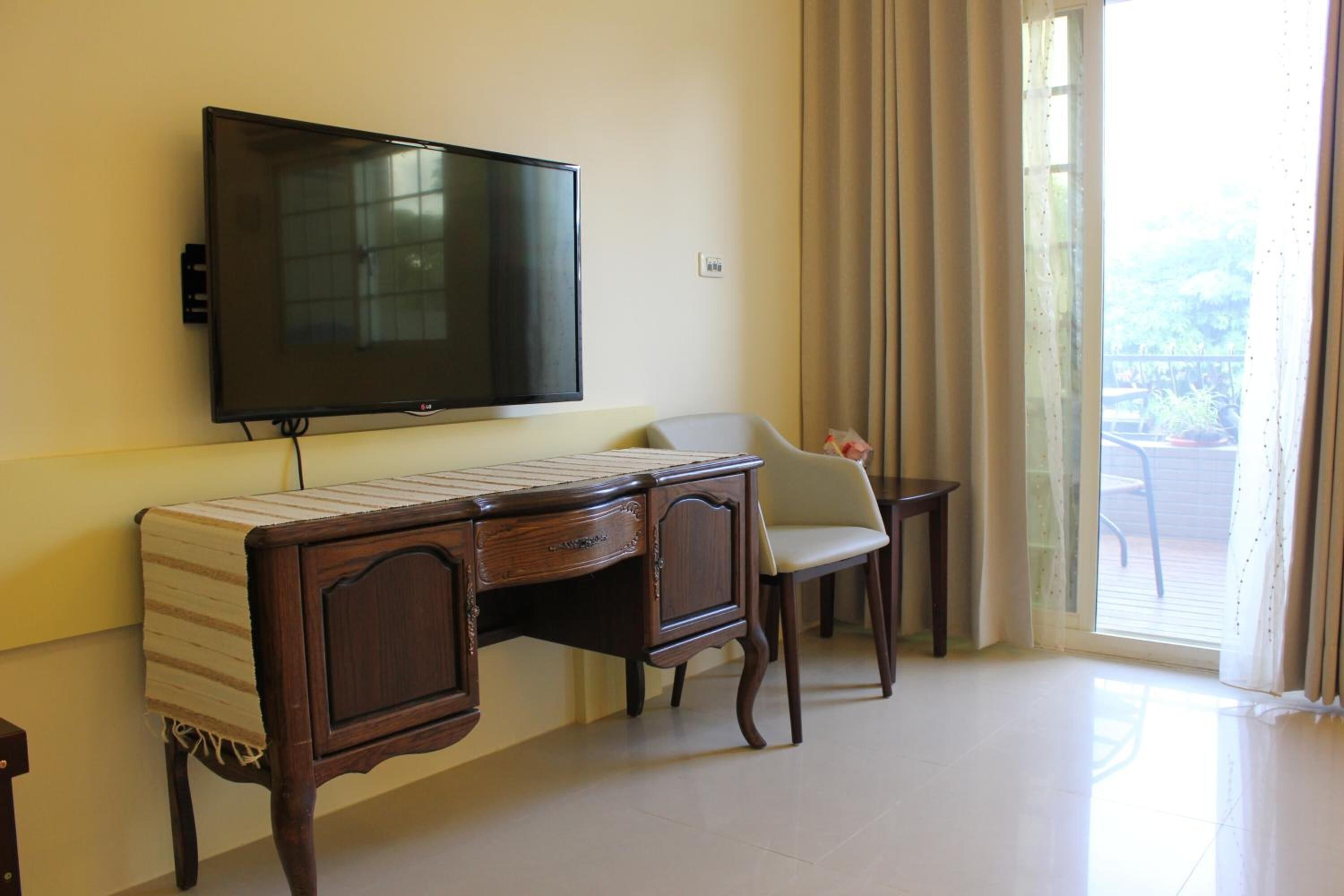 A hotel room with a flat-screen TV mounted on the wall, a wooden desk with a striped tablecloth, a chair, and a view of the outdoors through a large window with curtains.
