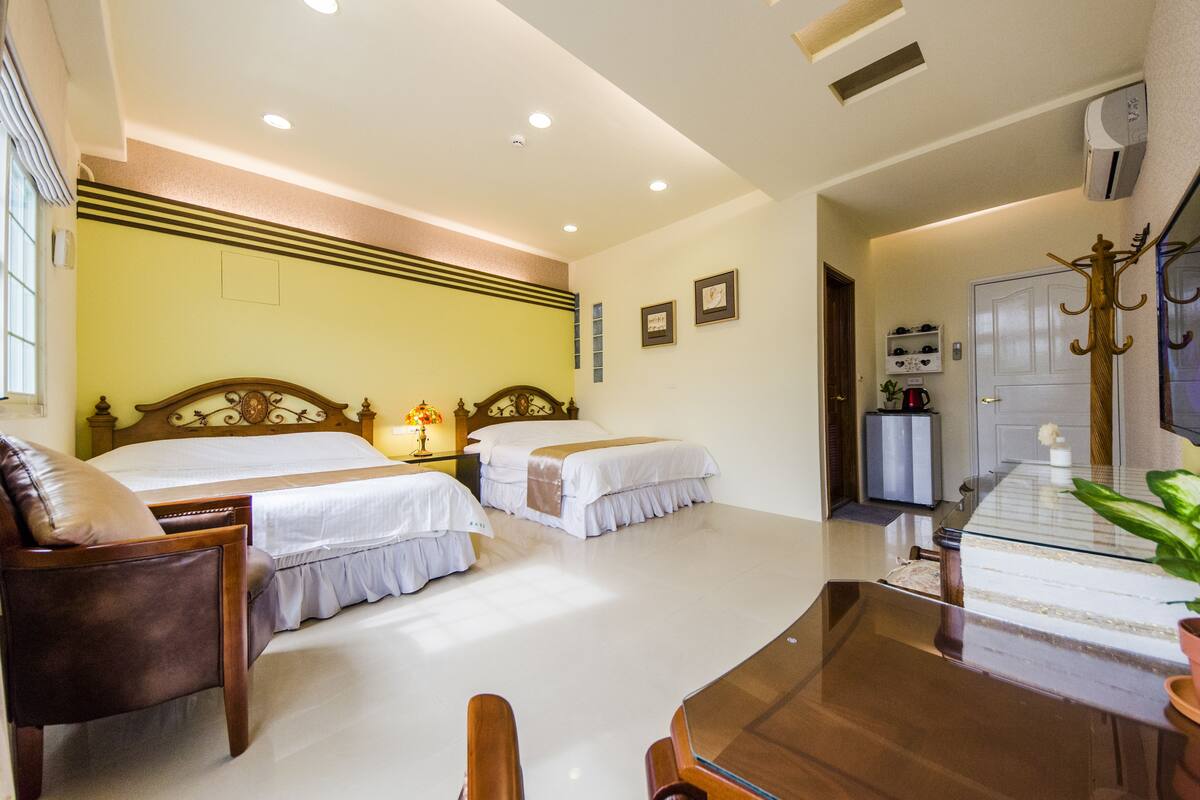 Quarto de hotel com duas camas, uma cadeira de madeira, uma mesa de vidro, um cabideiro e uma televisão.