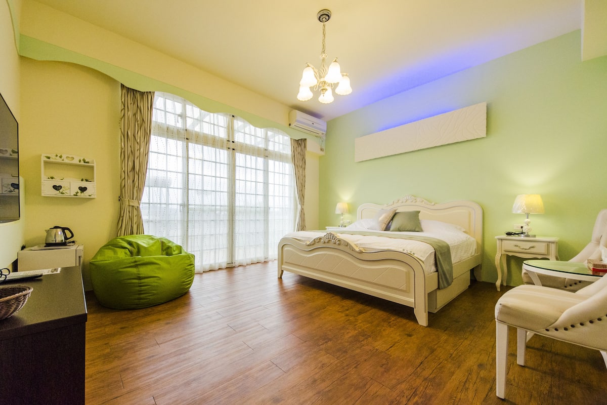 Quarto com cama, mesas de cabeceira, uma cadeira, um bean bag e uma janela com cortinas.