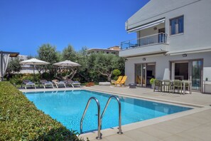 A modern outdoor pool area with lounge chairs and umbrellas, surrounded by greenery and a two-story building. - Sunlight Breeze Villa (Chania)