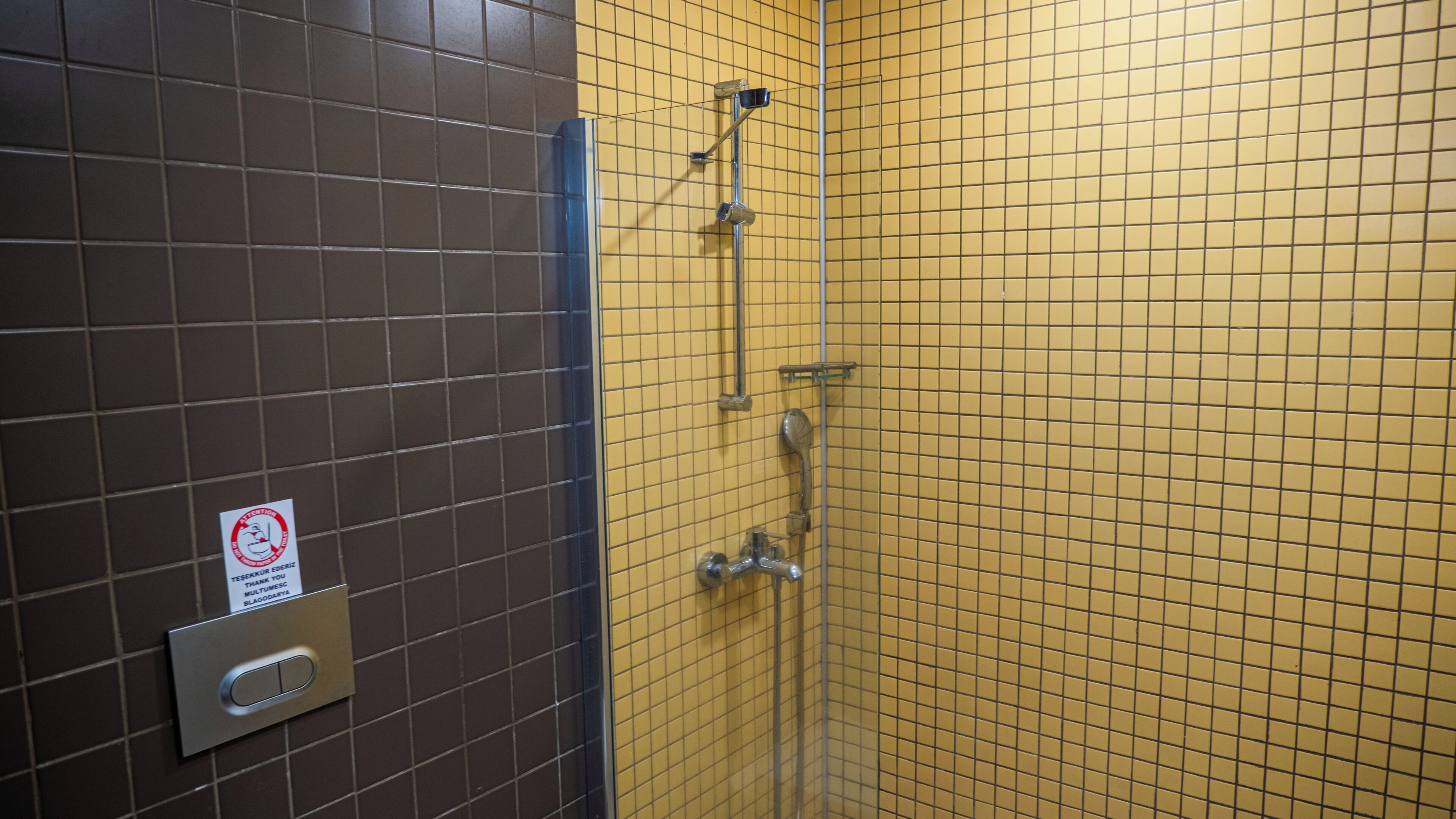 A bathroom with brown and yellow tiled walls, a hand dryer, and a sign with a red circle and white background.