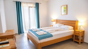 Una cama bien tendida con sábanas blancas y una manta azul, una mesita de noche con una lámpara, una mesita auxiliar de madera, un escritorio con un control remoto y una ventana con cortinas azules.